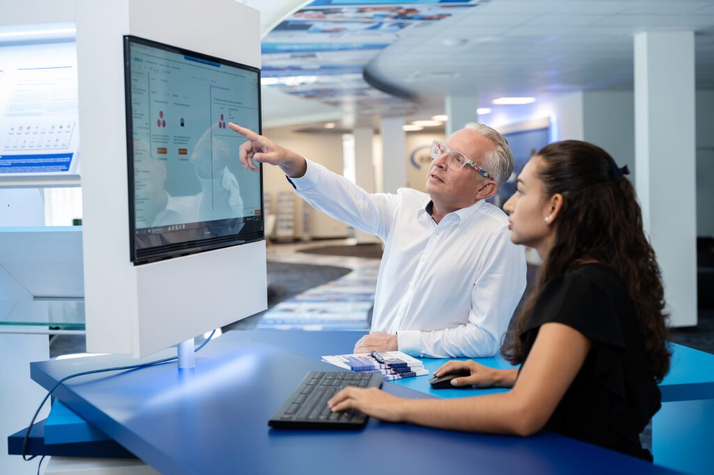 Eine weibliche Mitarbeiterin und ein männlicher Mitarbeiter der Consulting4IT stehen vor einem Bildschirm in der Academy. Der Mann zweigt der Frau etwas auf dem Bildschirm, während sie Maus uns Tastatur bedient.