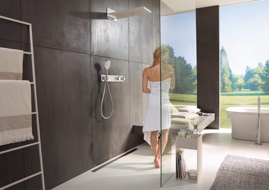 Ein modernes, helles Badezimmer mit Dusche und Badewanne der Firma hansgrohe sowie einem großen, bodentiefen Fenster. Eine blonde Frau, die ein Handtuch um ihren Körper trägt, verlässt gerade die Dusche.