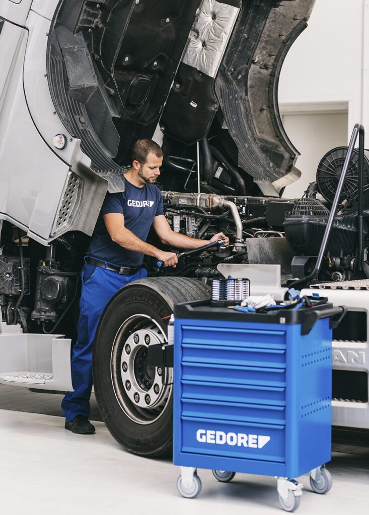 Werkzeugrollwagen der Firma Gedore. Im Hintergrund ein Mechaniker mit Gedore-T-Shirt, der an einem Lastwagen arbeitet.