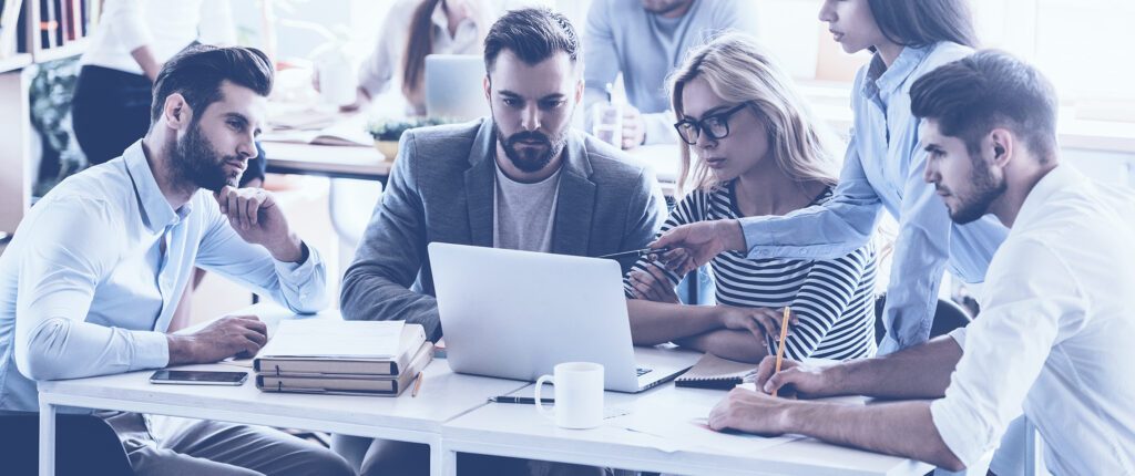 Fünf Personen im Fokus, die gemeinsam an einem Tisch sitzen oder stehen und konzentriert auf den Bildschirm eines Laptops schauen und teilweise deuten. Tassen, Unterlagen und Stifte auf dem Tisch, im Hintergrund weitere Tische und Personen unscharf zu erkennen.