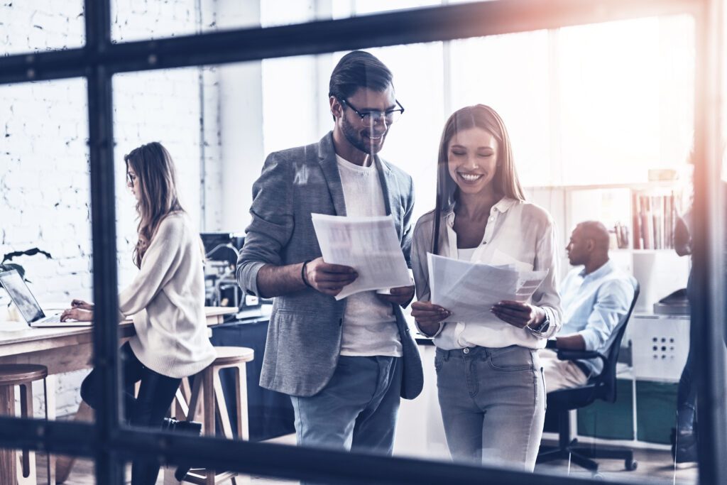 Ein Mann und eine Frau in Business Casual Kleidung, die lächelnd nebeneinander stehen und auf die Ausdrucke in ihrer beider Hände schauen. Im Hintergrund unscharf zwei weitere Personen an Schreibtischen. Bild aufgenommen durch eine Glasfassade mit dunklen Streben.