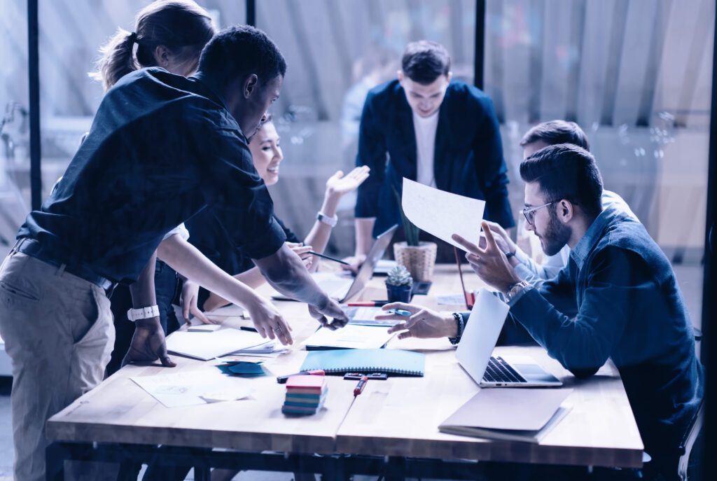 6 Personen in Business-Kleidung, die teils sitzend, teils vorgebeugt stehend an einem Besprechungstisch angeregt miteinander diskutieren, gestikulieren und dabei teilweise Blätter und Stifte in der Hand haben. Auf dem Tisch Laptops, Papiere, Hefte, Post-its und Stifte.