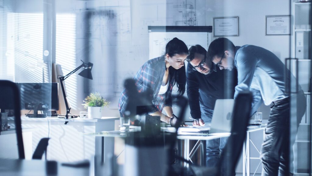 Zwei Männer und eine Frau in einem hellen Büro, aufgenommen durch eine Glasscheibe und verschiedene Spiegelungen, die sich mit konzentrierten Gesichtern über einen Tisch beugen, auf dem ein Laptop und Unterlagen stehen bzw. liegen.