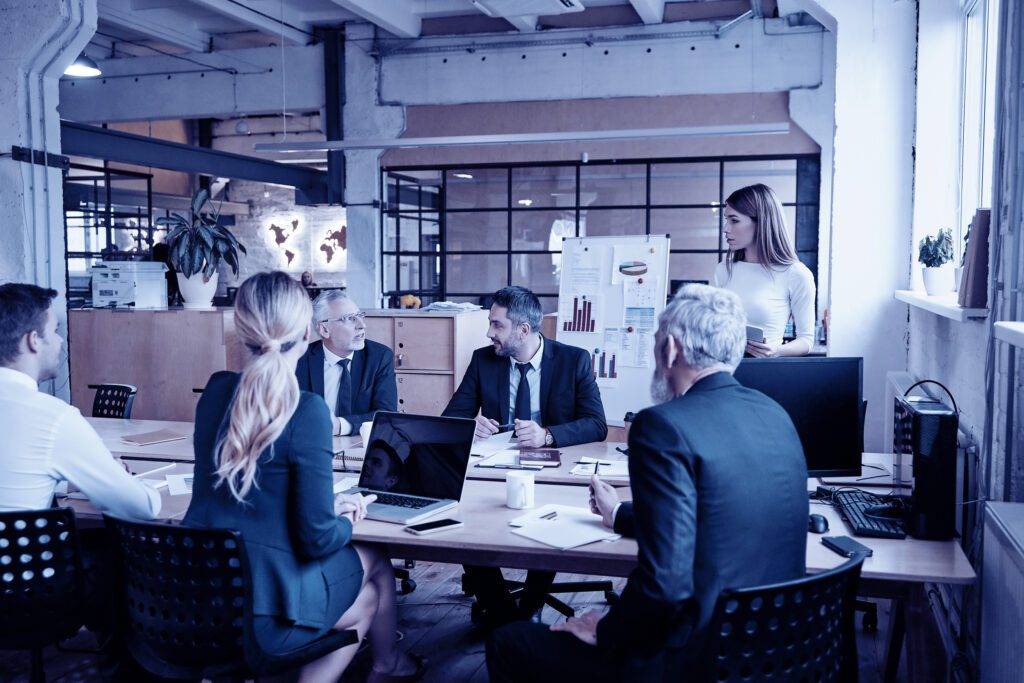 Eine große, offen gestaltete Bürofläche im Indurstriedesign mit viel Glas, Stahl und Beton, zentral im Vordergrund ein Besprechungstisch mit sehr förmlich gekleideten Personen - in Anzug mit Krawatte oder Kostüm - mit Laptops und Unterlagen und einer Flipchart mit Diagrammen im Hintergrund.
