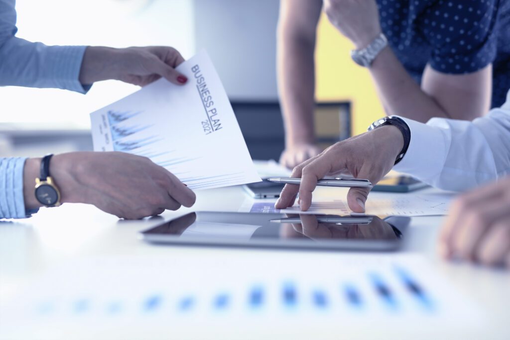 Ausschnitt, auf dem die Hände und Arme von drei Personen zu sehen sind. Eine Frau hält ein Blatt mit der Aufschrift 'Business Plan 2021' und Statistikdiagrammen in der Hand. Ein Mann hat die Finger auf einem weiteren Ausdruck auf dem Tisch gespreizt, einen Kugelschreiber zwischen den Fingern, daneben liegt ein Tablett, das so schräg seitlich aufgenommen wurde, dass man den Bildschirm nur als spieglende, schwarze Oberfläche sieht. Im Hintergrund sieht man eine dritte Person, die sich mit Hand und Ellbogen auf den Tisch stützt.