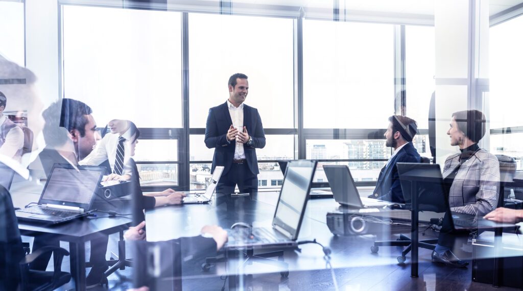 Eine Meetingsituation, aufgenommen durch eine leicht spiegelnde Scheibe. Ein Mann im Anzug steht hinter einem Besprechungstisch, mit gestikulierenden Händen, offenbar etwas erklärend, weitere Menschen sitzen am Tisch, vor ihnen Laptops und schauen ihn lächelnd an.