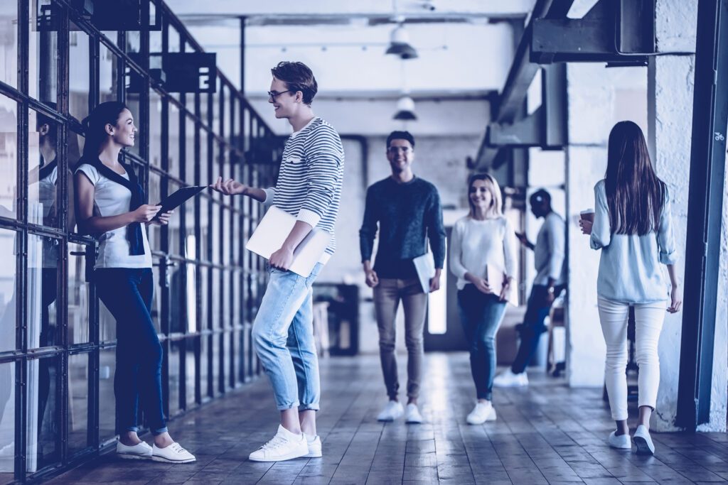 Ein heller offener Gang. Mehrere Menschen laufen aneinander vorbei zwei unterhalten sich.