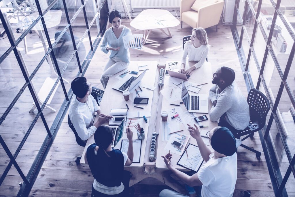 Ansicht einer Konferenzszene von schräg oben - sechs Personen an einem Konferenztisch, eine Frau sitzt halb schräg auf dem Tisch und präsentiert lächelnd einen Ausdruck mit einem Diagramm. Auf dem Tisch Laptops, Unterlagen und diverse Büroutensilien.