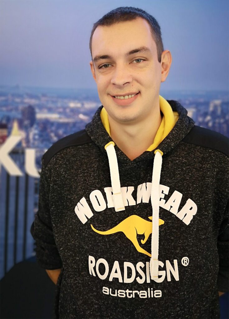 Mann mit kurzen Harren und einem grauen Hoodie lächelt schüchtern in die Kamera.