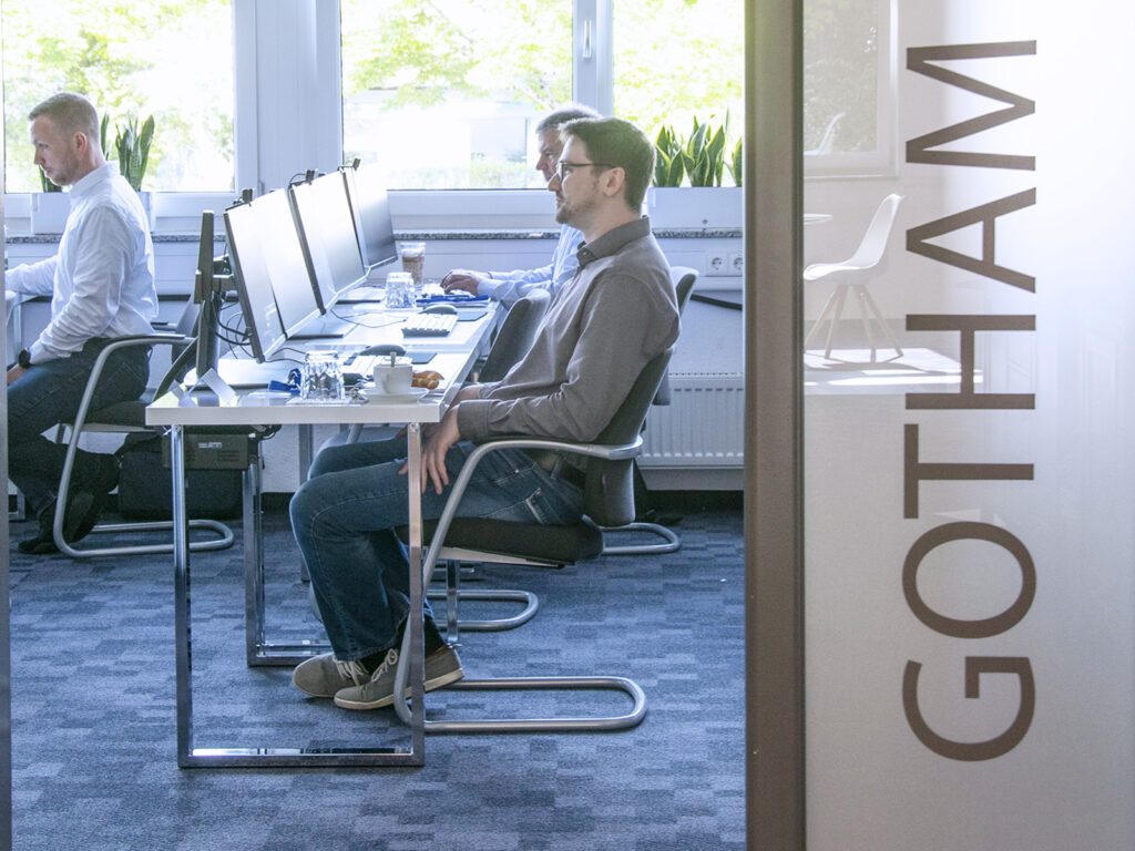 Blick in einen Academy Schulungssraum. Zwei Teilnehmer und der Trainer sind zu sehen. Außerdem kann man den Namen des Schulungsraumes "Gotham" an der Tür lesen.