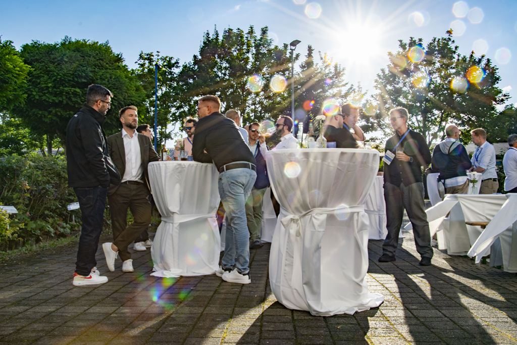 Stehtische auf einem Hof oder Parkplatz, die mit weißen Hussen versehen sind und an denen einige Menschen in hellem Sonnenschein stehen, Wein und Cocktails trinken und sich unterhalten, während am Himmel über ihnen eine sehr helle und strahlende Sonne steht und das Bild mit gleißenden Strahlen versieht.