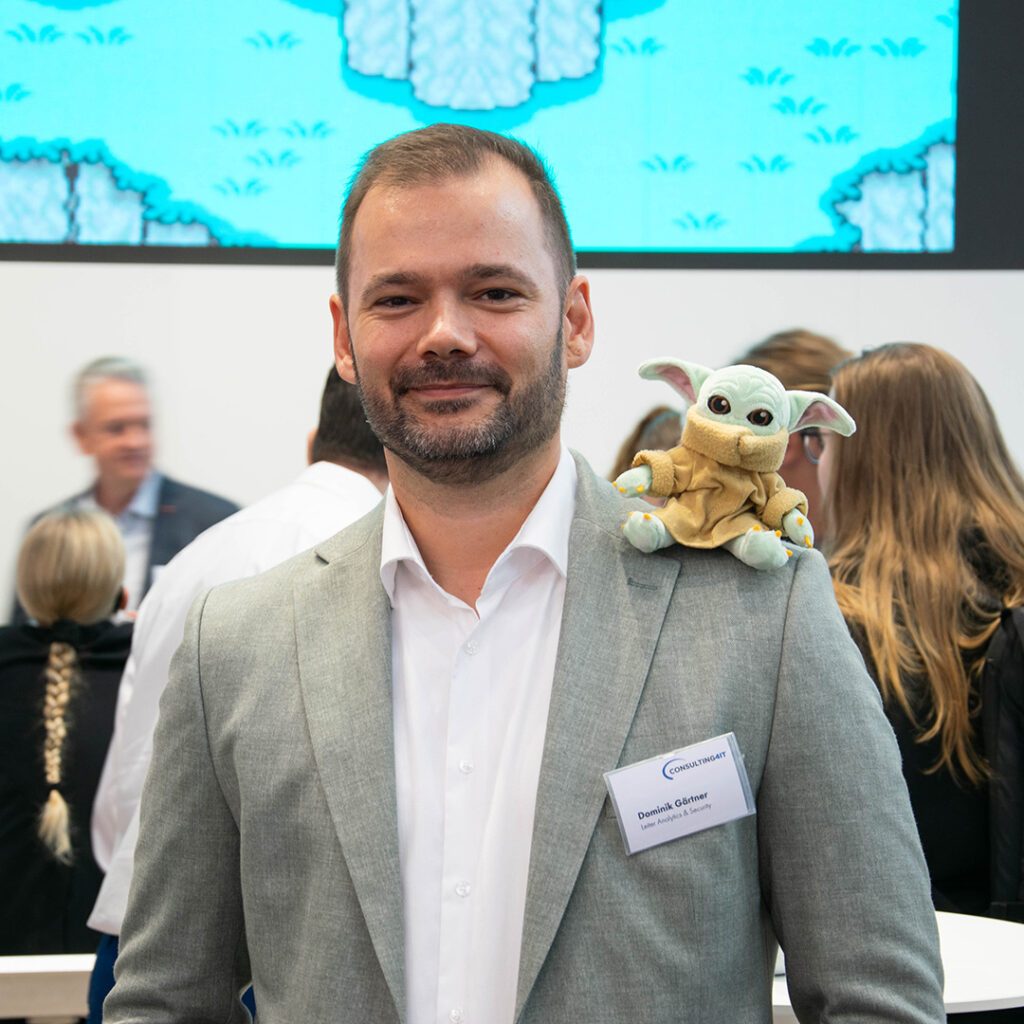 IT Analytics Leiter Dominik Gärtner, lächelnd, in weißem Hemd und grauem Jacket, der eine Plüschfigur von Miniyoda scherzhaft auf der Schulter sitzen hat, im Hintergrund weitere Personen.