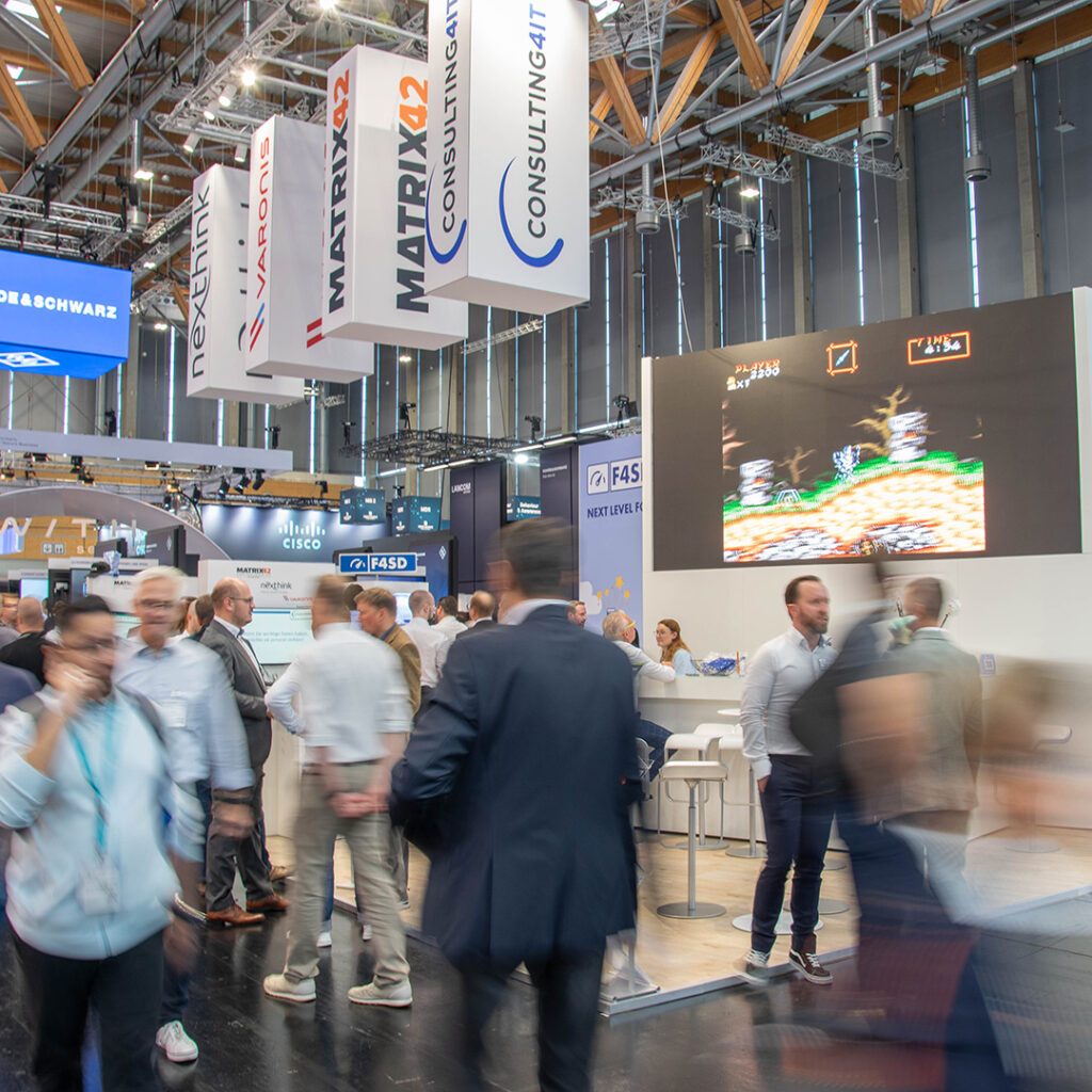 Ansicht von in Bewegungsunschärfe stark verwischten menschlichen Schemen, die am Consulting4IT Messestand vorbeieilen, auf dem Messestand verschiedene Personen, die scharf abgebildet sind.