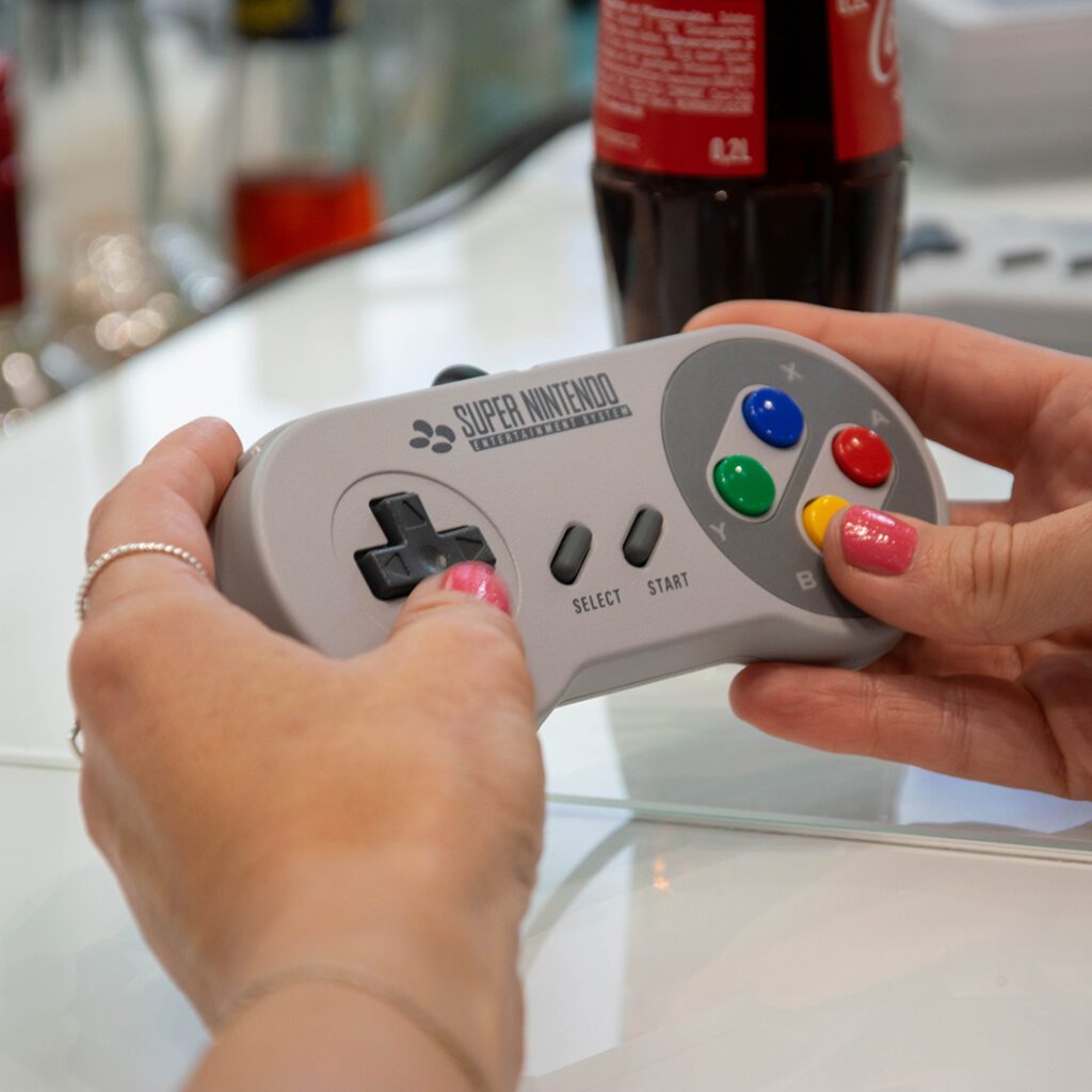 Zwei Frauenhände mit pink lackierten Nägeln, die den Controller einer Supernintendo bedienen.