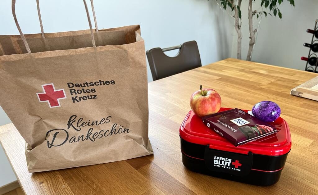 Eine braune Papiertüte, eine Brotdose, eine Tafel Schokolade, ein Apfel und ein lila Osterei vom Deutschen Roten Kreuz als kleine Aufmerksamkeit nach der Blutspende.