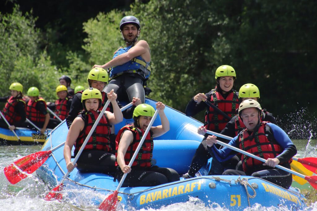 Consulting4IT-Mitarbeiter beim Rafting in einem blauen Schlauchboot mit roten Schwimmwesten und grünen Helmen.
