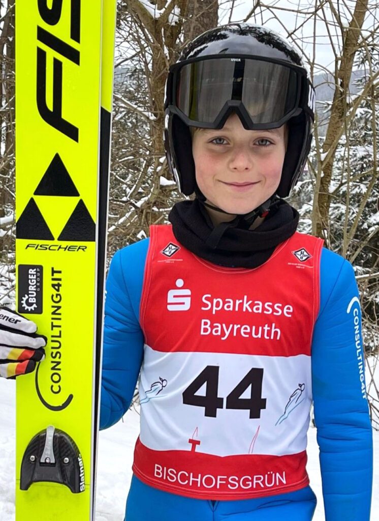 Julian Schonhardt in Ski-Outfit, mit Helm und Ski in der Hand vor winterlicher Kulisse.
