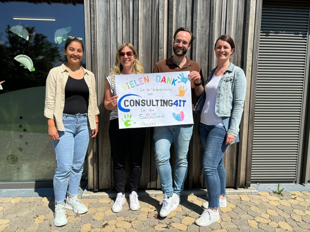 Patrick Hild, Linda Schmittner und zwei verantwortliche Damen des Kindergartens Don Bosco vor einer Holzwand im Freien bei Sonnenschein, während sie ein Dankeplakat der Kinder in den Händen halten.