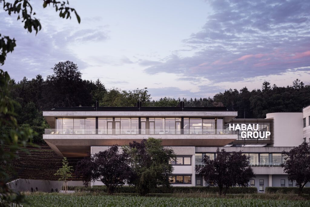 Ansicht des Firmengebäudes der Habau Group im Dämmerlicht inmitten einer grünen Anlage und Wald, überspannt von einem nahezu wolkenlosen blauvioletten Himmel.