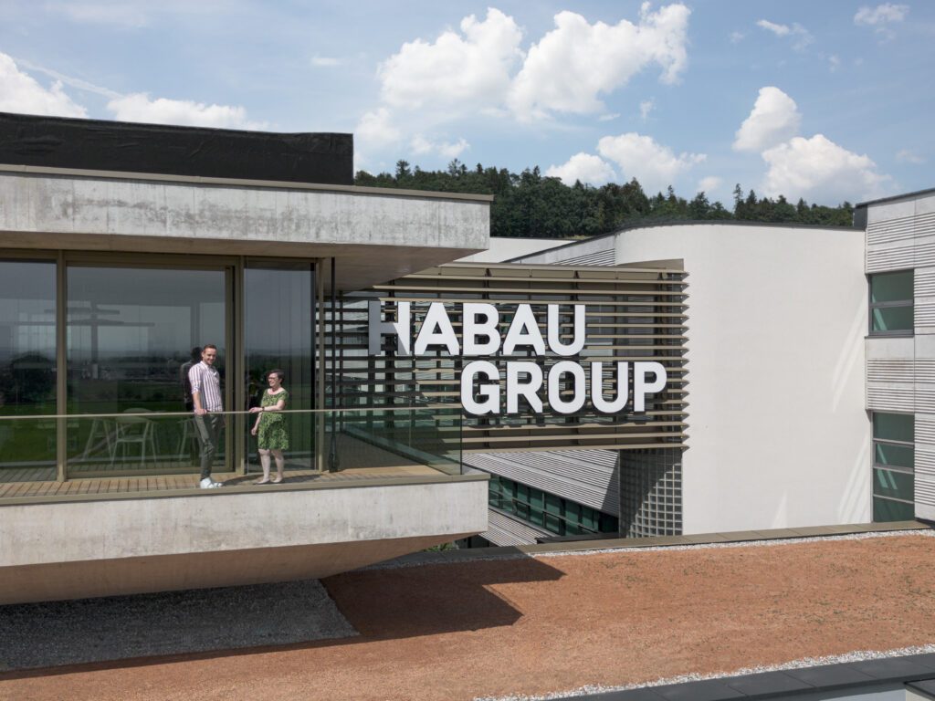 Ansicht der Firmenzentrale der Habau Group in Österreich mit Firmenschriftzug und zwei Personen auf einem Balkon..