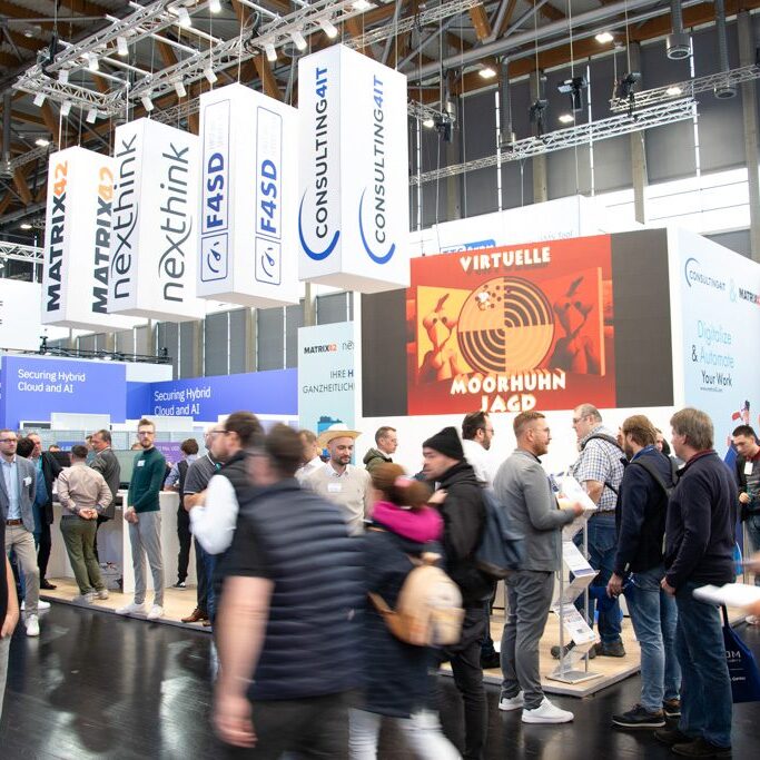Messestand der Consulting4IT auf der it-sa in Nürnberg mit zahlreichen Besuchern. Über dem Stand hängen Würfel mit den Logos „Matrix42“, „Nexthink“, „F4SD“ und „Consulting4IT“. Im Hintergrund ist ein großer Bildschirm mit der Aufschrift „Virtuelle Moorhuhn-Jagd“ zu sehen.