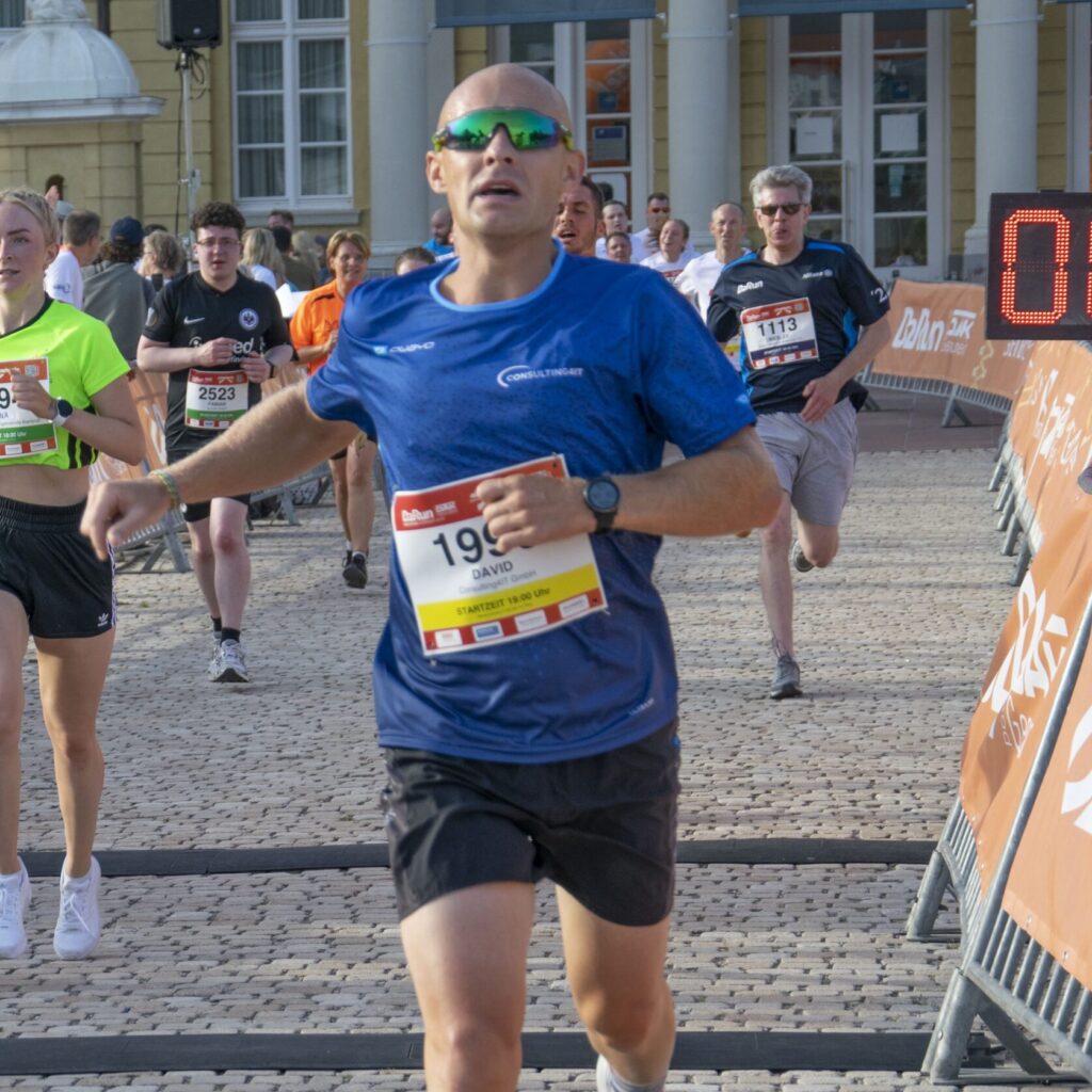 Consulting4IT Mitarbeiter David Hajduk beim Zieleinlauf des B2Run Karlsruhe 2022. Er trägt ein blaues Laufshirt, schwarze Shorts und eine verspiegelte Sonnenbrille.