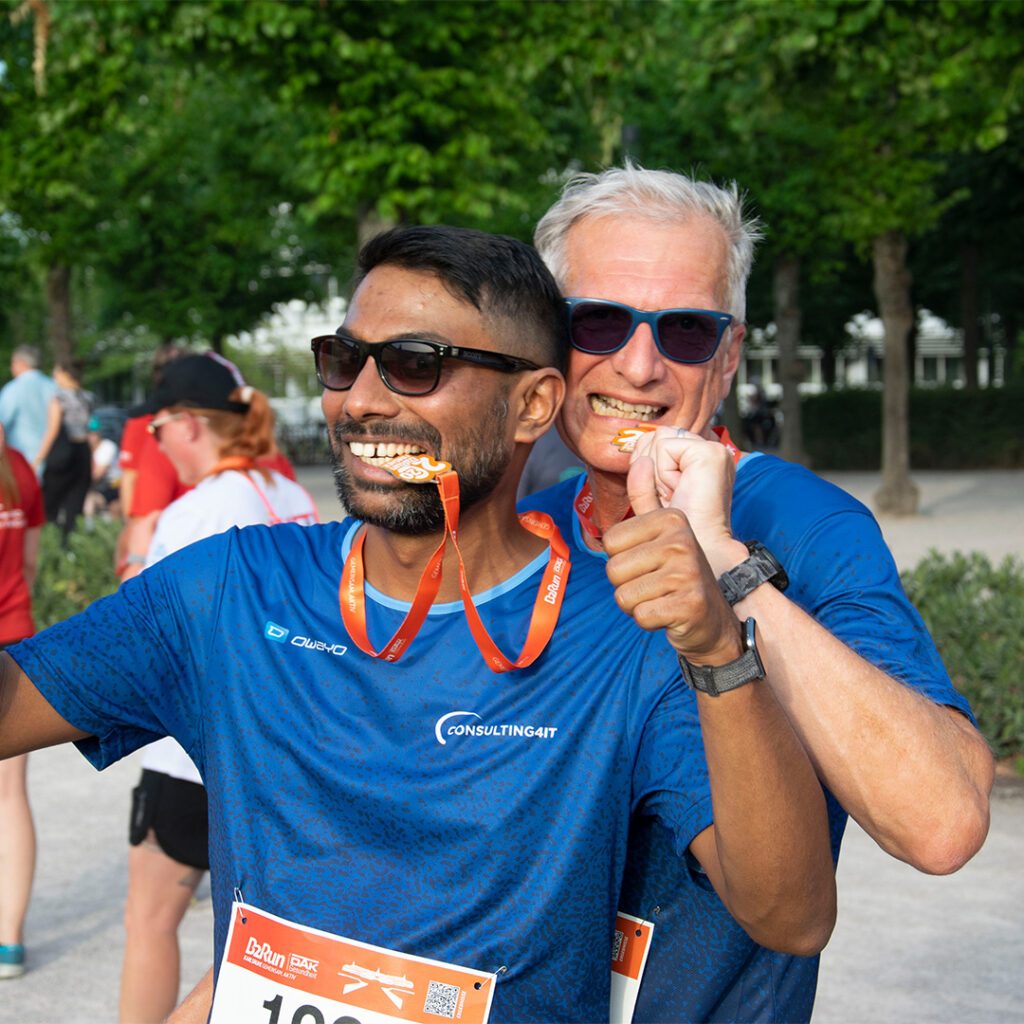 Zwei Männer in blauen Laufshirts feiern nach dem B2Run und beißen lächelnd in ihre Medaillen, die an orangefarbenen Bändern hängen. Beide tragen Sonnenbrillen und strahlen Freude aus. Im Hintergrund sind andere Läufer und grüne Bäume zu sehen.