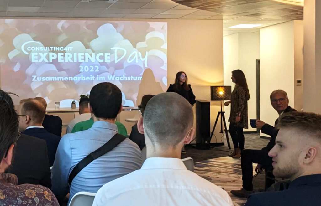 Ein Veranstaltungsraum mit Teilnehmern, die in Stuhlreihen sitzen und auf eine beleuchtete Leinwand blicken. Auf der Leinwand steht „Consulting4IT Experience Day 2022 – Zusammenarbeit im Wachstum“.