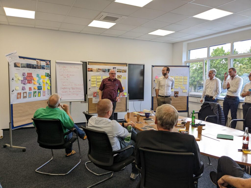 Ein Workshop mit mehreren Teilnehmern in einem Konferenzraum. Aufgestellte Pinnwände zeigen bunte Haftnotizen, Schaubilder und Notizen. Zwei Männer moderieren die Sitzung, während andere am Tisch sitzen oder an der Seite stehen und zuhören.