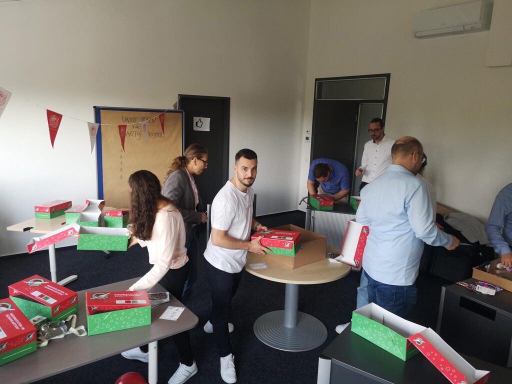 Ein Team von Mitarbeitern füllt Geschenkkartons an mehreren Tischen in einem Konferenzraum für die Aktion "Weihnachten im Schuhkarton".