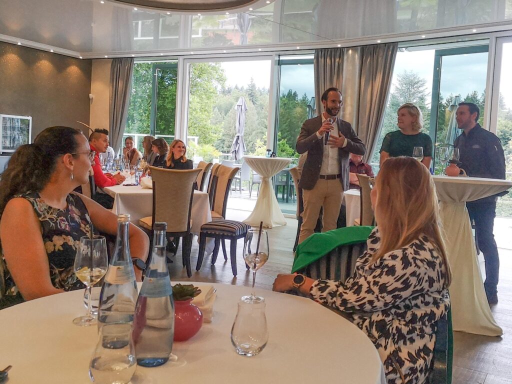 Feier in einem Raum mit Glasfronten. Ein Mann hält eine Rede mit einem Mikrofon, während die Gäste an Tischen sitzen. Im Hintergrund stehen zwei Personen an einem Stehtisch, und die Einrichtung wirkt stilvoll und modern.