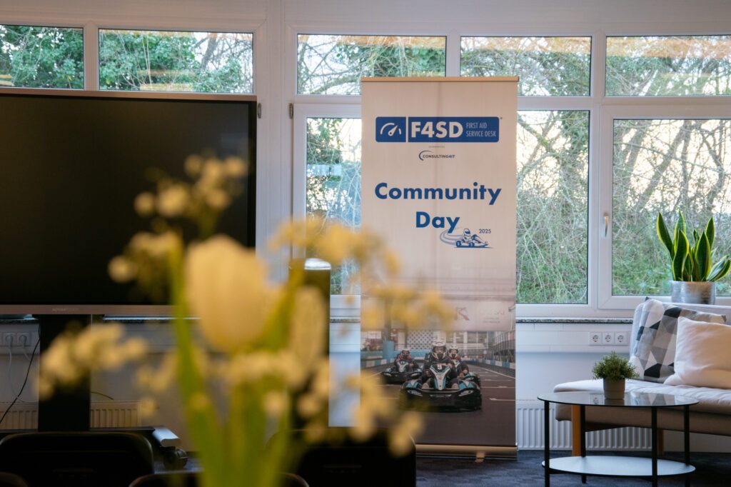 Ein heller Konferenzraum mit einem großen Bildschirm und einem aufgestellten Banner für den „F4SD Community Day 2025“. Im Vordergrund sind unscharfe weiße Blumen, während der Raum mit Pflanzen und Sitzmöbeln gemütlich dekoriert ist.