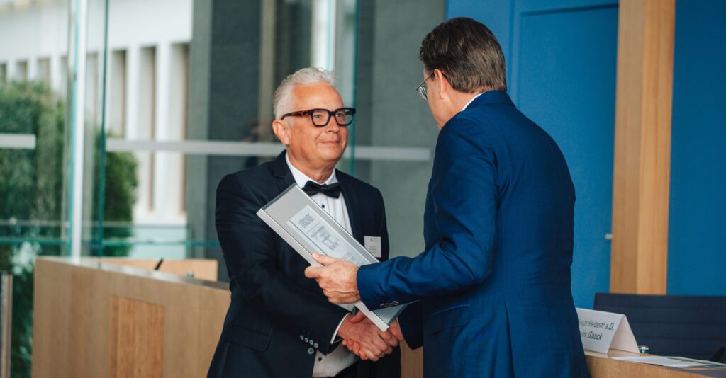 Ein Mann überreicht Mirko Oesterhaus, Geschäftsführer der Consulting4IT GmbH, bei einer offiziellen Veranstaltung die Urkunde zum Senator.