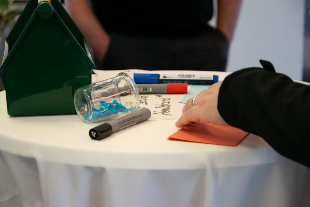 Nahaufnahme eines runden Tisches mit Moderationsmaterialien wie Stiften, Notizzetteln und Pinnnadeln. Eine Hand greift gerade nach einer orangenen Karte, daneben steht ein grünes Kästchen – im Hintergrund unscharf eine stehende Person.