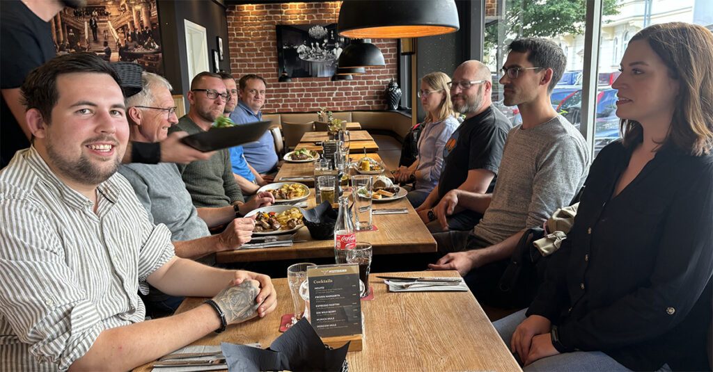 Mehrere Menschen sitzen an Tischen in einem Restaurant. Einige haben Teller mit Essen und Getränken vor sich und lächeln dabei in die Kamera. Ein Kellner überreicht gerade ein weiteres Gericht.