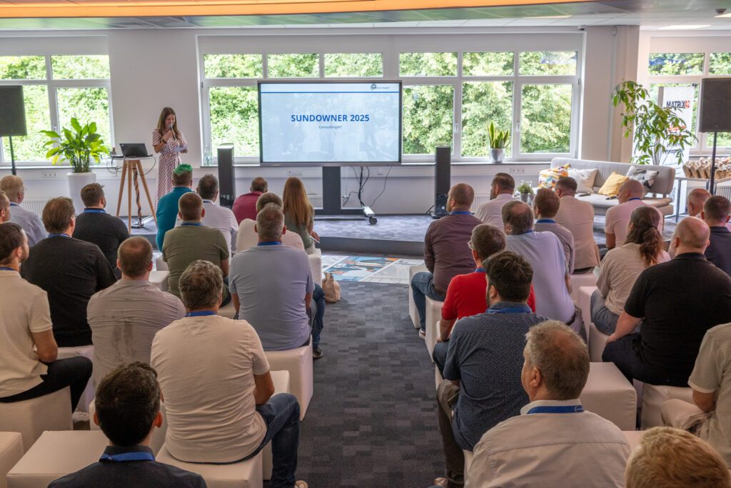 Eine Frau spricht auf einer Bühne vor einem Publikum, während auf dem Bildschirm „Sundowner 2025 – Consulting4IT“ angezeigt wird.