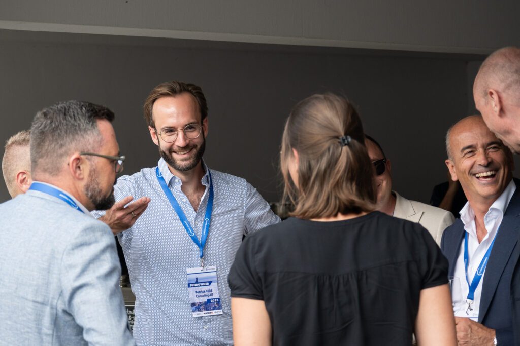 Gruppe von Menschen im Gespräch bei einer Veranstaltung, ein Mann mit Namensschild „Sundowner 2025“ lacht.