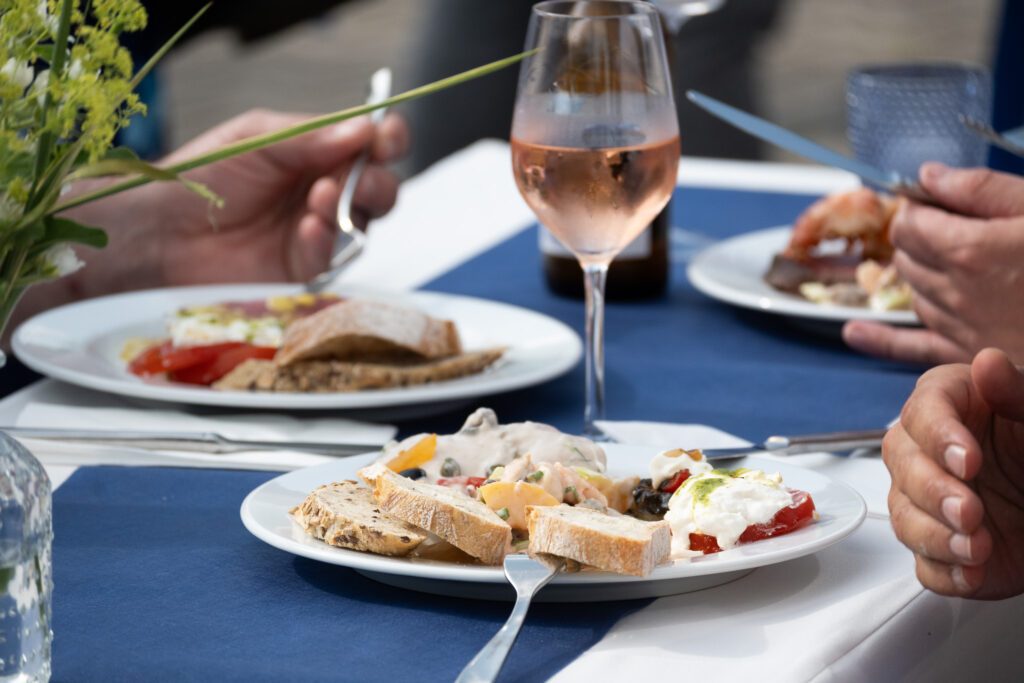 Mehrere Personen essen gemeinsam an einem Tisch, im Fokus ein Teller mit Baguette, Meeresfrüchtesalat und Mozzarella, daneben ein Glas Rosé.