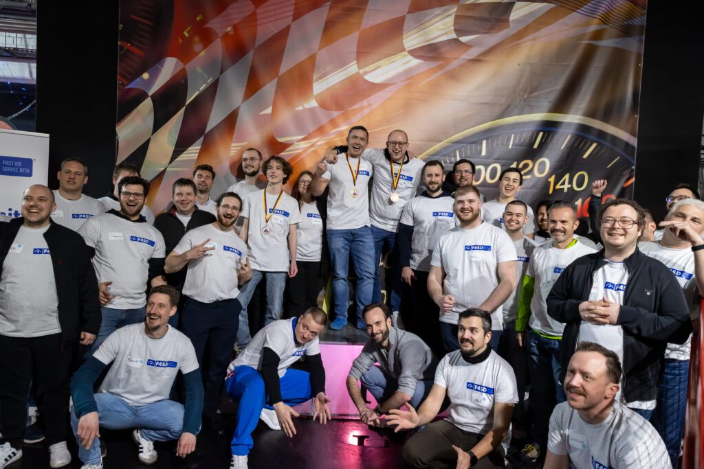 Group photo from the F4SD Community Day with about 30 participants. In the center of the photo, three winners stand on a podium wearing gold, silver, and bronze medals. Everyone in the photo is wearing F4SD T-shirts; many are laughing, cheering, or making gestures of joy.